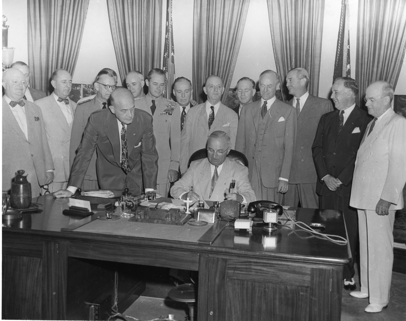 President Harry S. Truman is seated at his desk in the Oval Office, signing the National Security Act Amendments of 1949, which converted the National Military Establishment into a new Department of Defense and made other changes in the national security system.