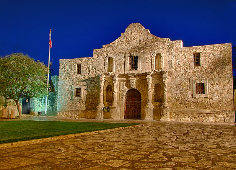 A picture of the Alamo at night with the Texas flag waving to the side. 